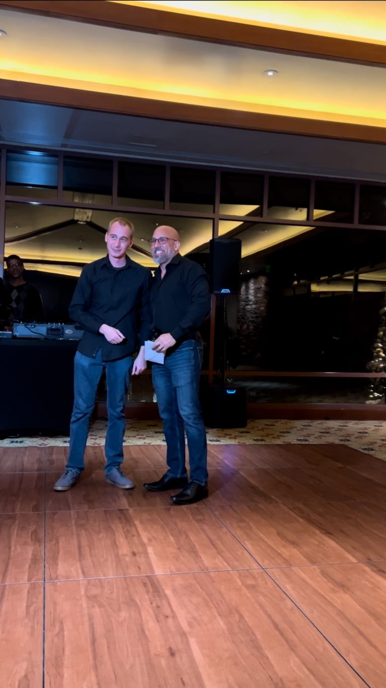 A second angle of CEO James Farias presenting the Most Resilient Team Member Award at Appreciation Financial's 2024 Holiday Party, with a DJ visible in the background.